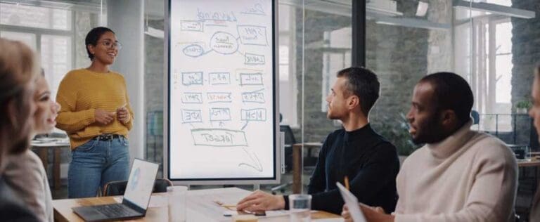 Project Manager Makes a Presentation for a Young Diverse Creative Team in Meeting Room in an Agency. Colleagues Sit Behind Conference Table and Discuss Business Development, User Interface and Design.