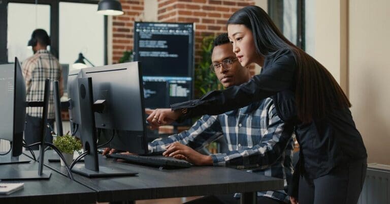 Two programers holding laptop with coding interface walking towards desk and sitting down talking about online cloud computing.