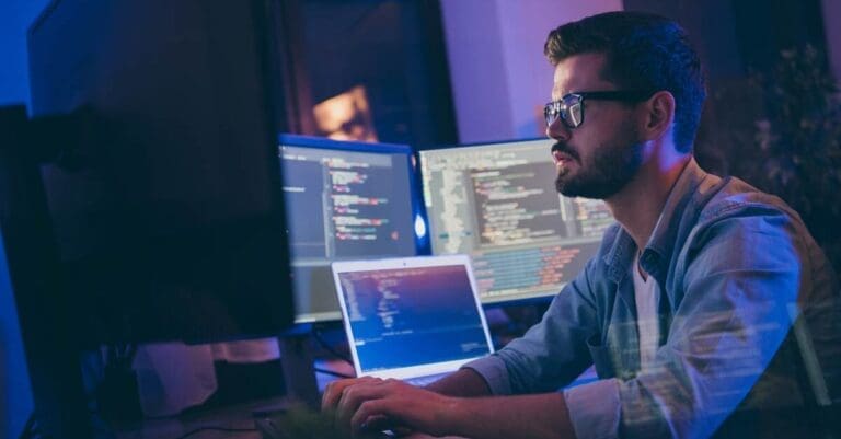 DevOps engineer in dark room looking at multiple computer screens, coding