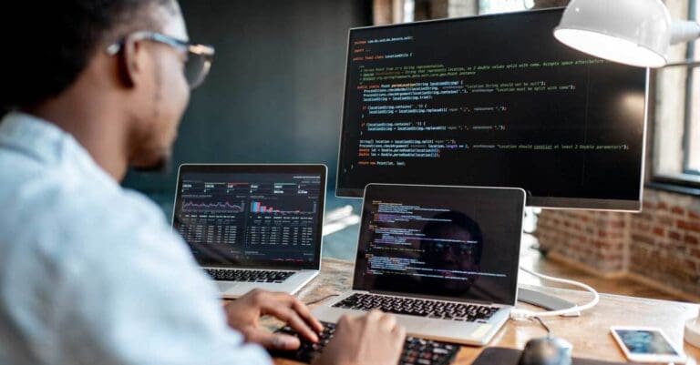 Young male programmer writing program code sitting at the workplace with three monitors in the office. Software development.