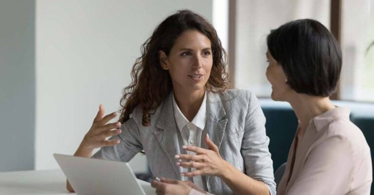 Two business women sit at desk in office discussing project details