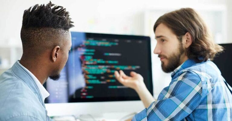 One of software developers pointing at coding on computer screen while explaining to his co-worker