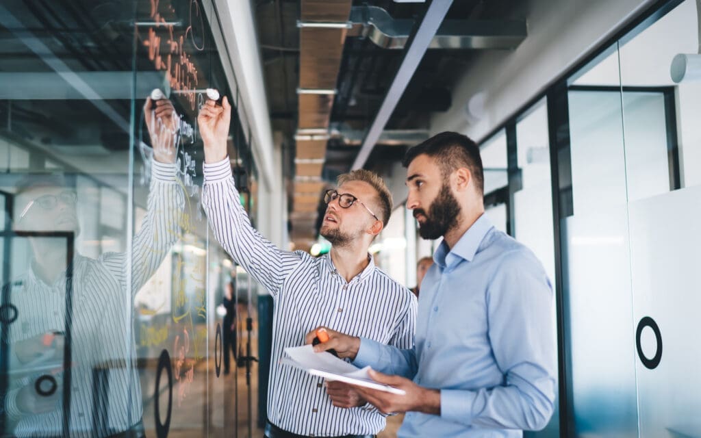 male employees cooperating on working process in business workspace, Caucasian men colleagues talking while planning and analyzing analytic from glass board during brainstorming meeting