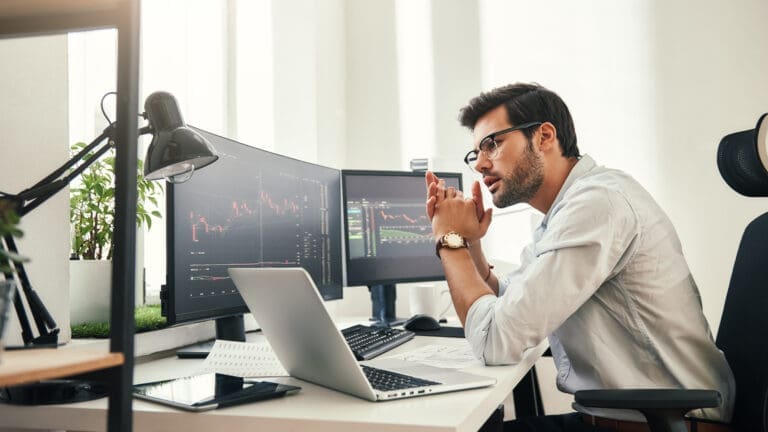 Trading strategy. Successful young trader in eyeglasses looking at analyzing trading charts on computer screens in while sitting in his modern office.